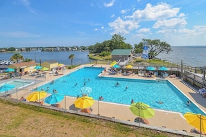 A large outdoor swimming pool with multiple lanes, surrounded by umbrellas and lounge chairs, with a view of the water and distant buildings. - Waterfront Retreat with Private Dock on the Sound (Kill Devil Hills)