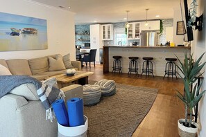 Una sala moderna con un sofá, otomanes y una mesa de centro. Hay una zona de cocina con armarios blancos y un taburete de bar.