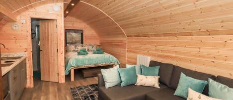 A cozy cabin interior with a wooden ceiling, a sofa with patterned cushions, a bed with a green blanket, and a small kitchen area.