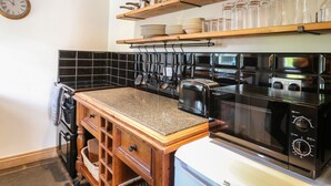 A kitchen with a wooden countertop, a black stove, and a microwave. There are shelves above holding dishes and glasses. - Hops Cottage (Ashbourne)