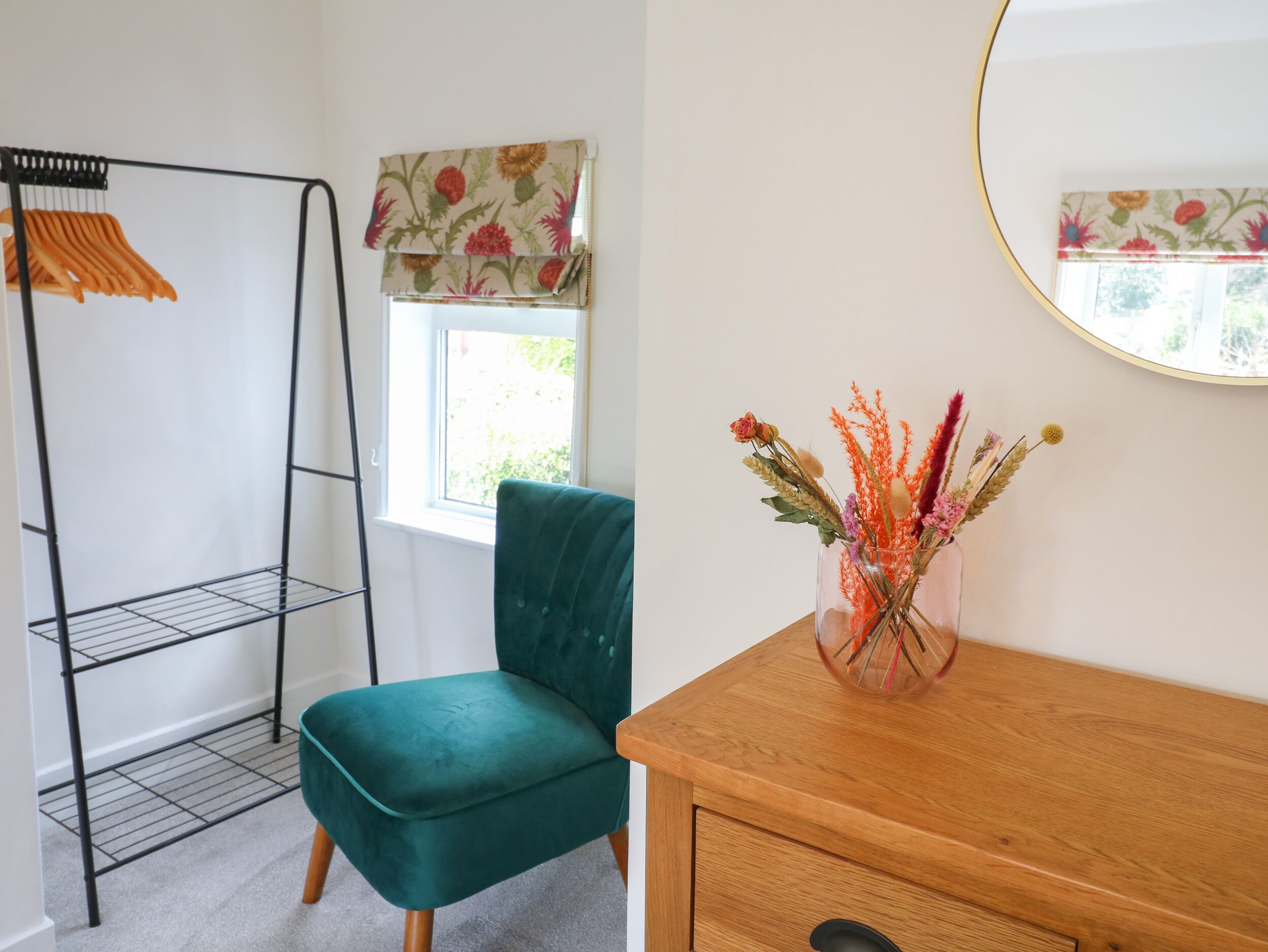 Une pièce comprenant un fauteuil de couleur turquoise, une commode en bois, un miroir et une fenêtre ornée de rideaux à motifs floraux.