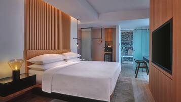 Une chambre d’hôtel moderne avec un grand lit, une télévision, une tête de lit en bois et, au fond, une petite kitchenette.