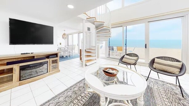 Un ampio soggiorno con una grande TV a schermo piatto, un tavolino da caffè bianco e posti a sedere confortevoli. Una scala a chiocciola conduce a un balcone con vista sull'oceano.