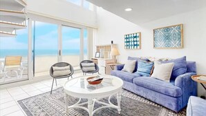 Un soggiorno moderno con un divano blu, un tavolino da caffè bianco e vista sulla spiaggia attraverso ampie vetrate scorrevoli.