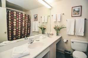 A bathroom with a large mirror, two sinks, white towels, a toilet, and a shower curtain.