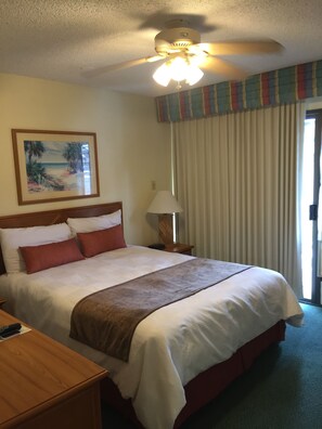 A hotel room with a bed, a nightstand, a lamp, a ceiling fan, a window with curtains, and a beach-themed painting on the wall.