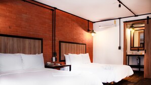 Ein Hotelzimmer mit zwei Betten, einem Holzkopfende, einer Ziegelwand, einem Holzboden, einem Nachttisch, einer Lampe und einem Badezimmer mit Spiegel und Waschbecken.