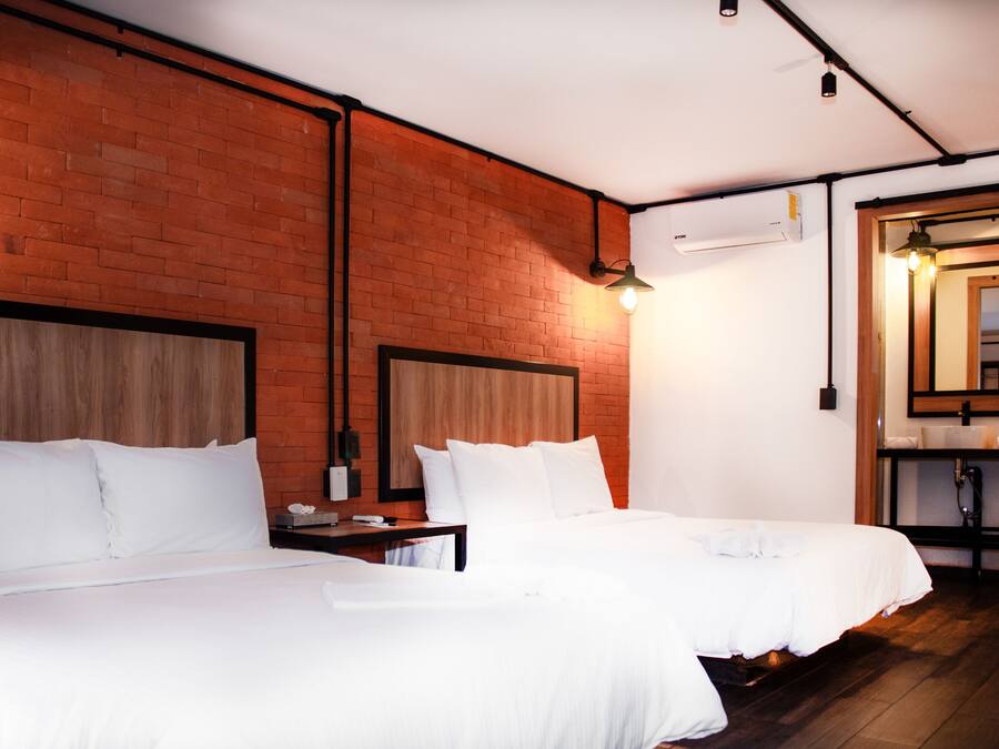 A hotel room with two beds, a wooden headboard, a brick wall, a wooden floor, a nightstand, a lamp, and a bathroom with a mirror and sink.