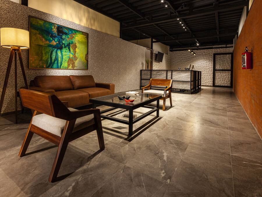 Um lobby moderno com um sofá de couro marrom, uma mesa de centro de vidro e uma cadeira de madeira. Há um abajur, uma pintura na parede e um extintor de incêndio fixado na parede.
