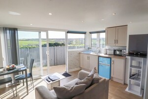 A modern kitchen with a blue refrigerator, a dining area with a table and chairs, and a large sliding glass door leading to a balcony with a view of the countryside.