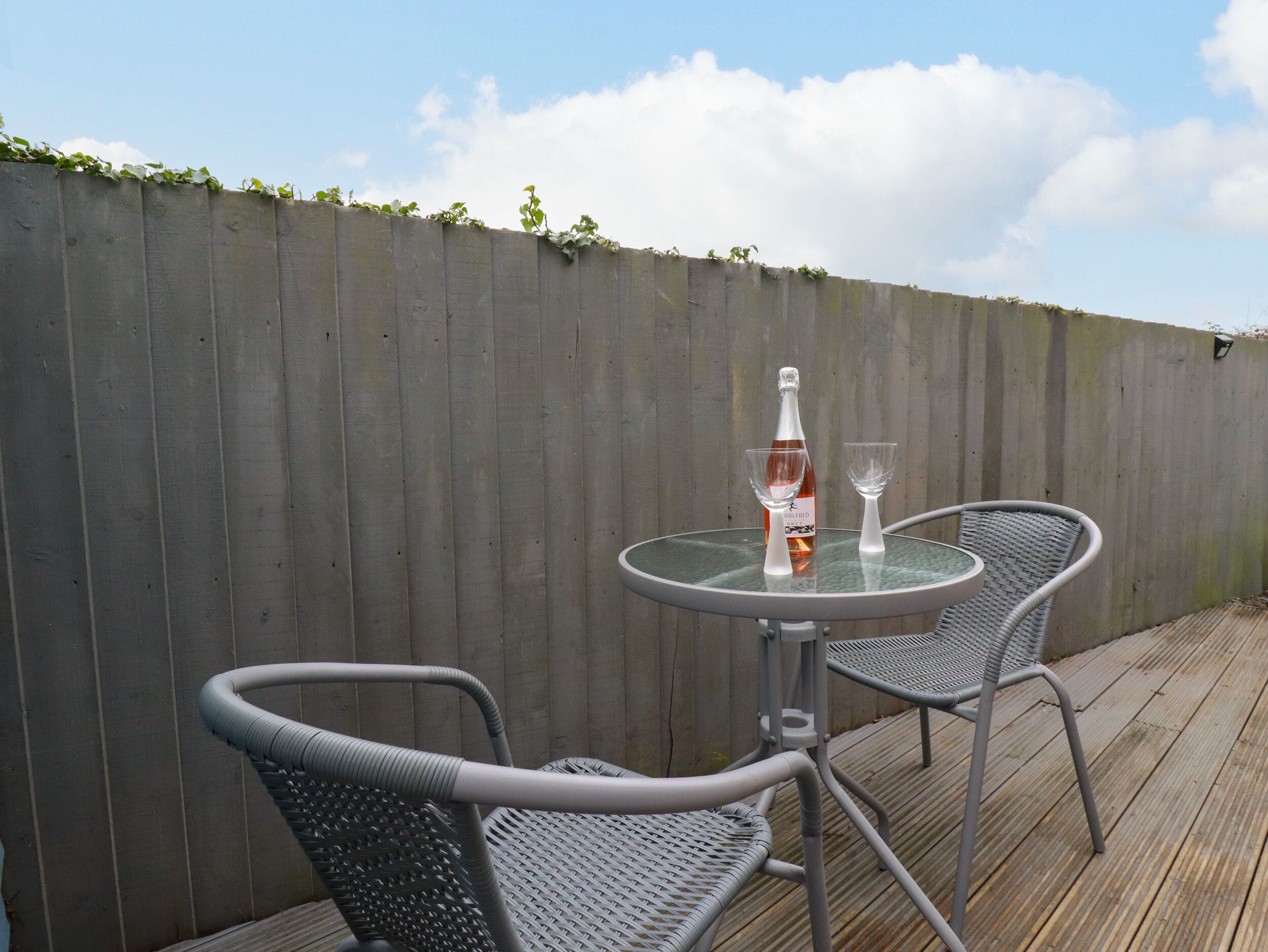 A round glass table with two metal chairs, a bottle of wine, and two wine glasses on a wooden deck.
