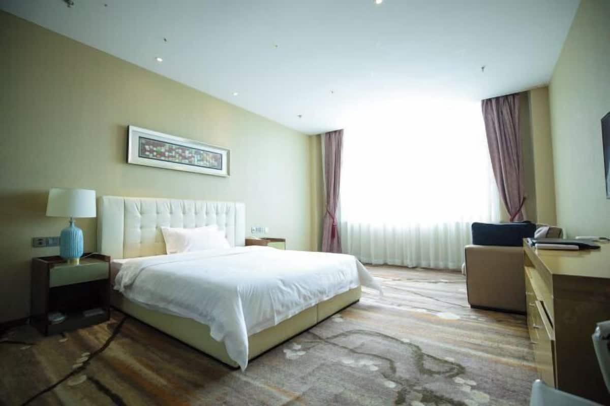 A hotel room with a large bed, a desk, and a chair. There is a window with curtains and a painting on the wall.