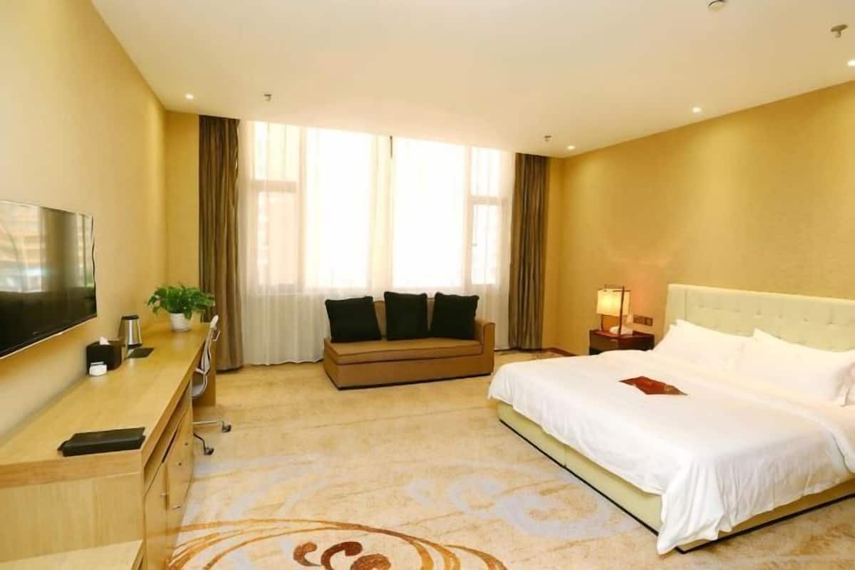 A hotel room with a bed, a sofa, a desk with a TV, and a window with curtains.