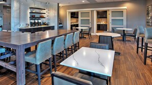 Un restaurante moderno con una mesa común larga, varias mesas más pequeñas y una zona de cocina con estantes y electrodomésticos.