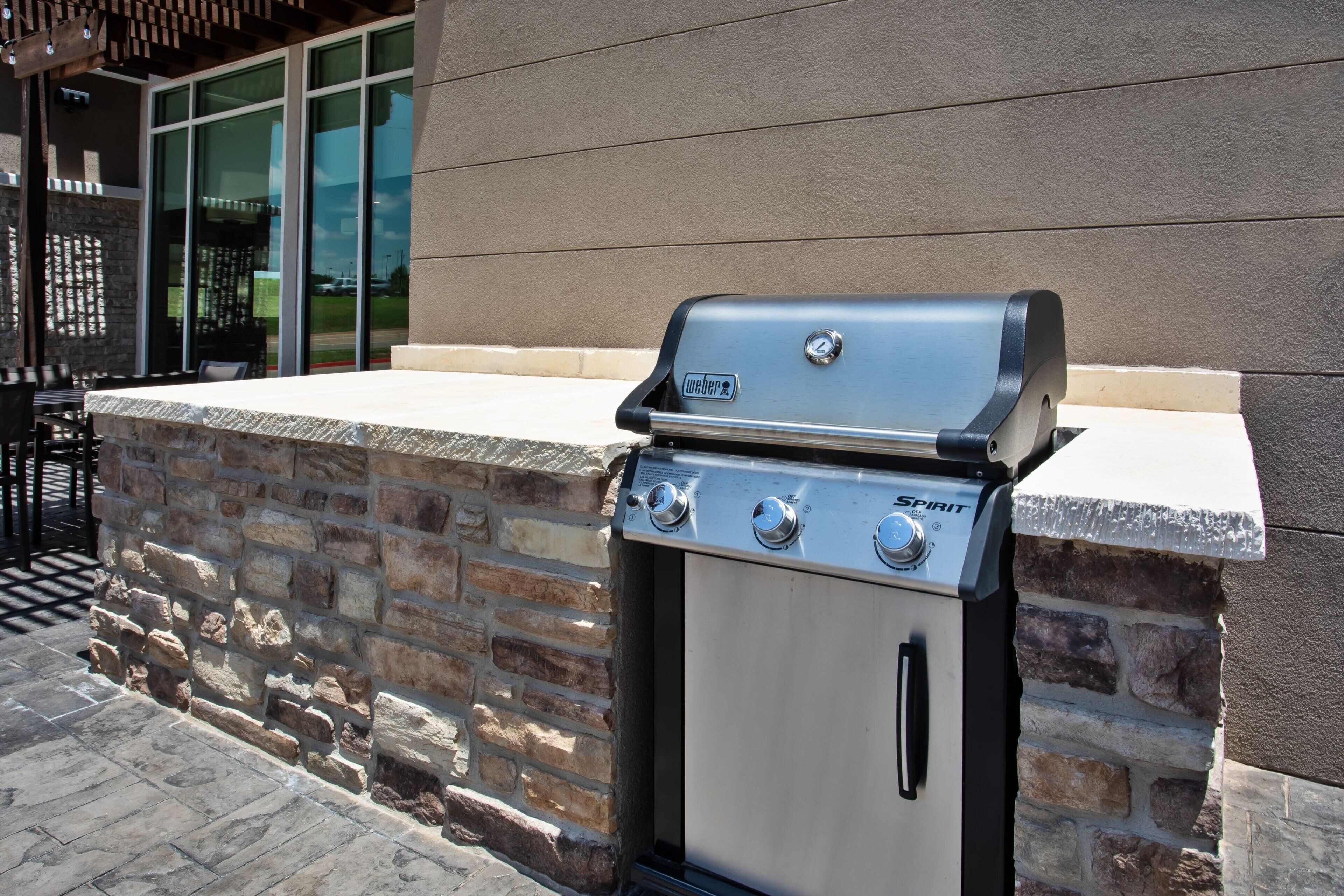 A Weber Spirit gas grill with a stone base, located outdoors near a building with large windows.