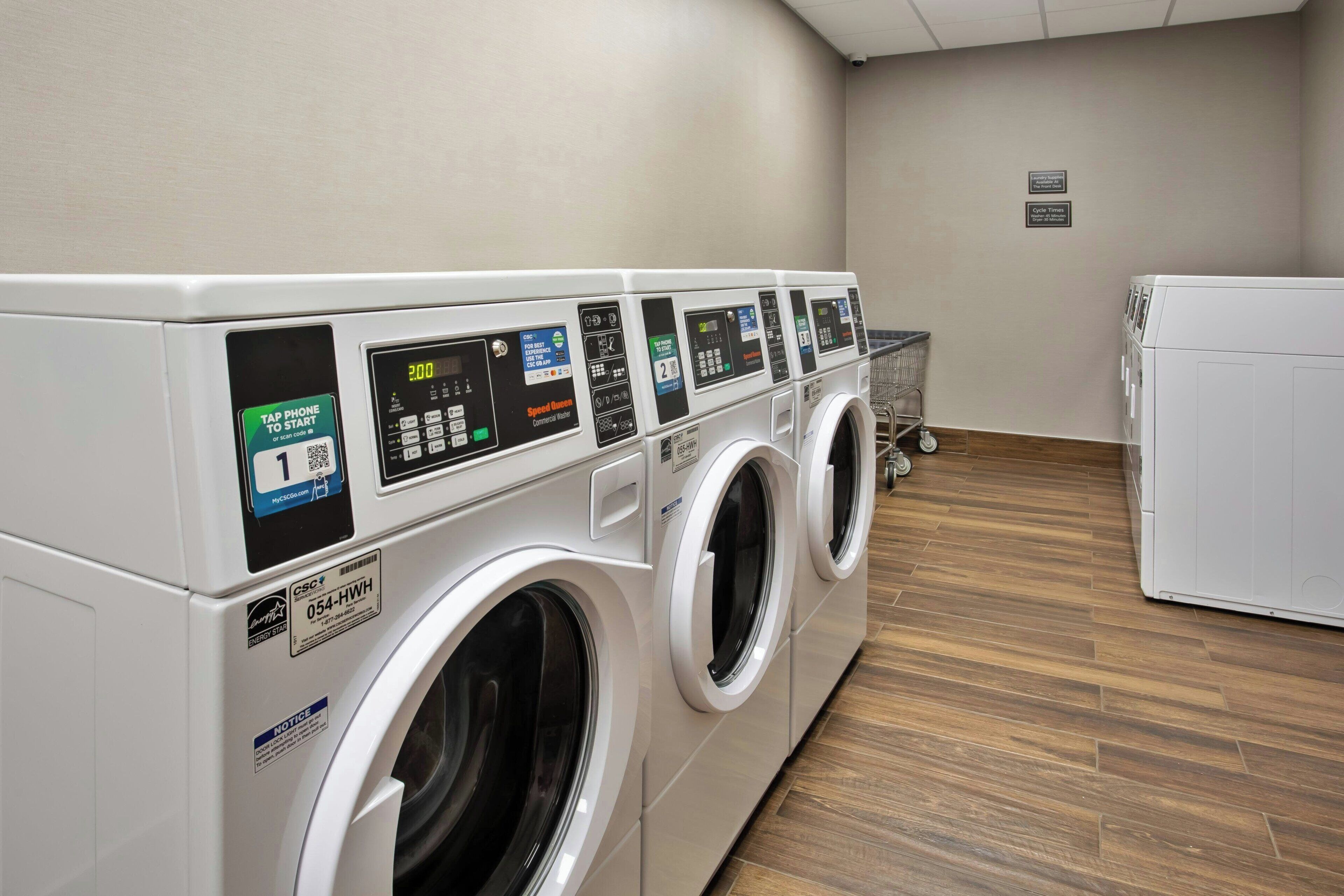 A row of front-loading washing machines in a laundry room with a wooden floor and a white refrigerator in the background.