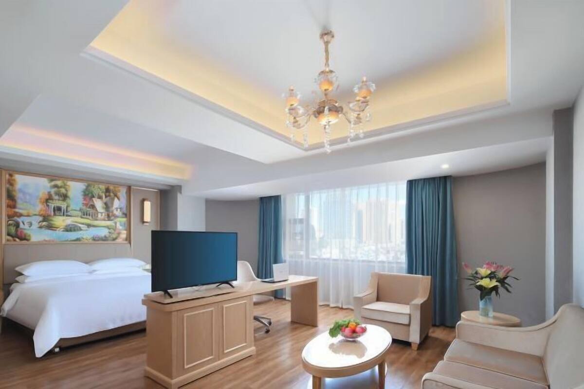 A hotel room with a bed, a TV, a desk, a chair, a sofa, and a painting on the wall.