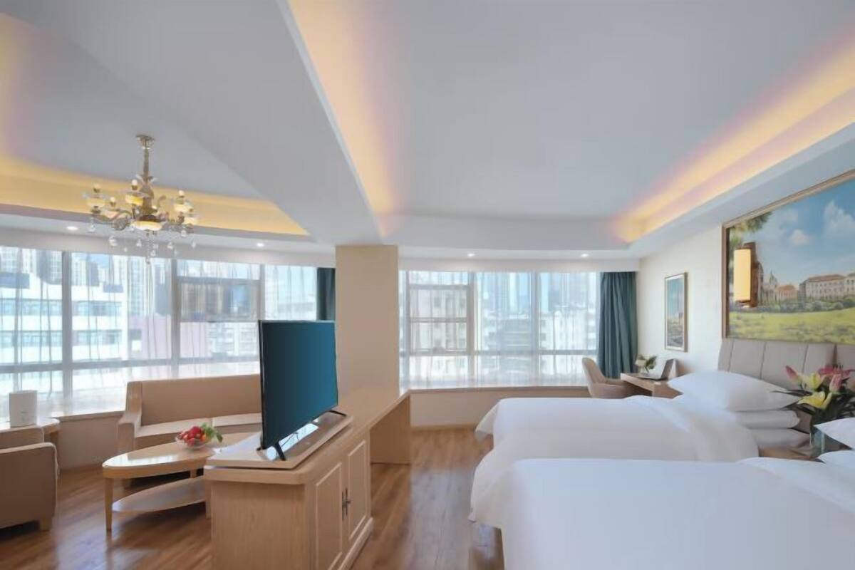 A hotel room with a large window, a desk with a computer, a TV, two beds, and a chandelier.