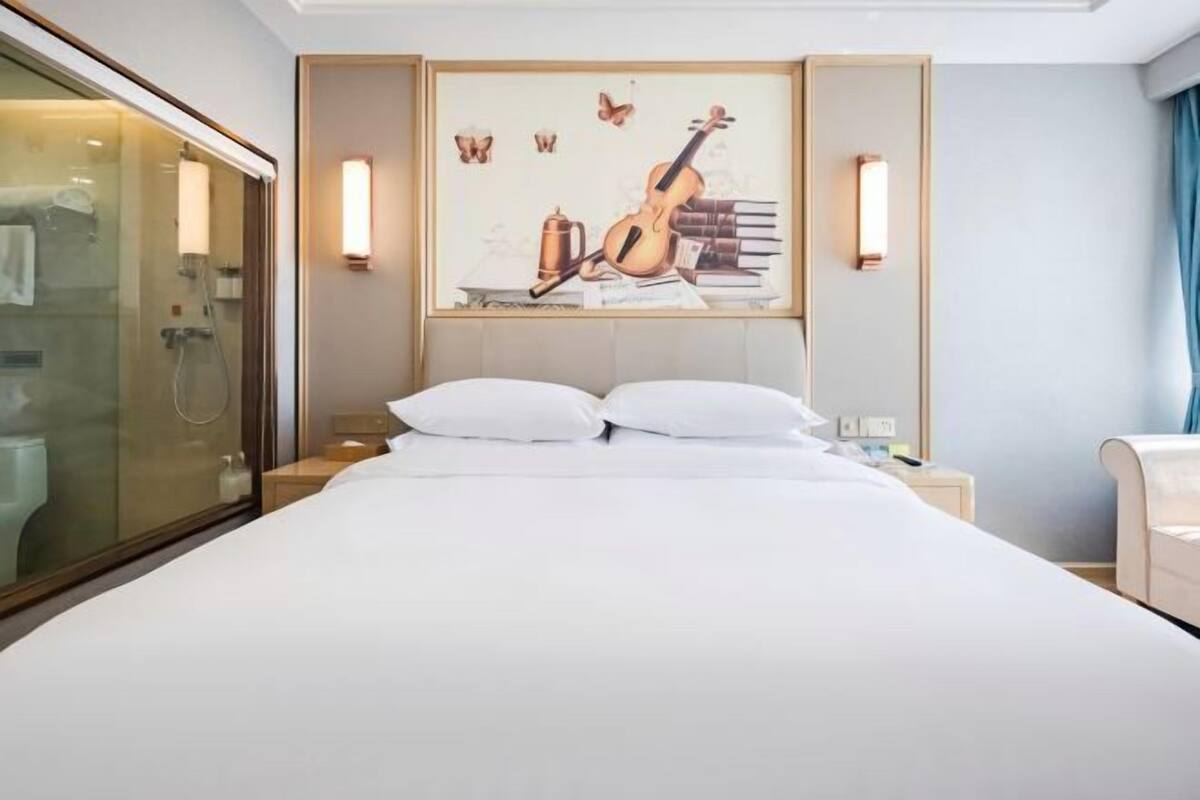 A hotel room with a bed, a glass shower enclosure, a painting of a violin, and a nightstand with a lamp.