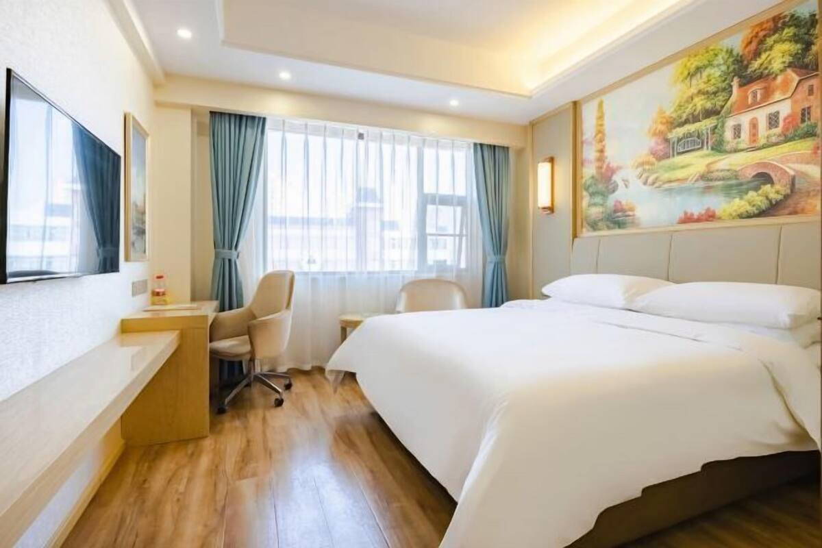 A hotel room with a bed, desk, chair, and a painting on the wall.