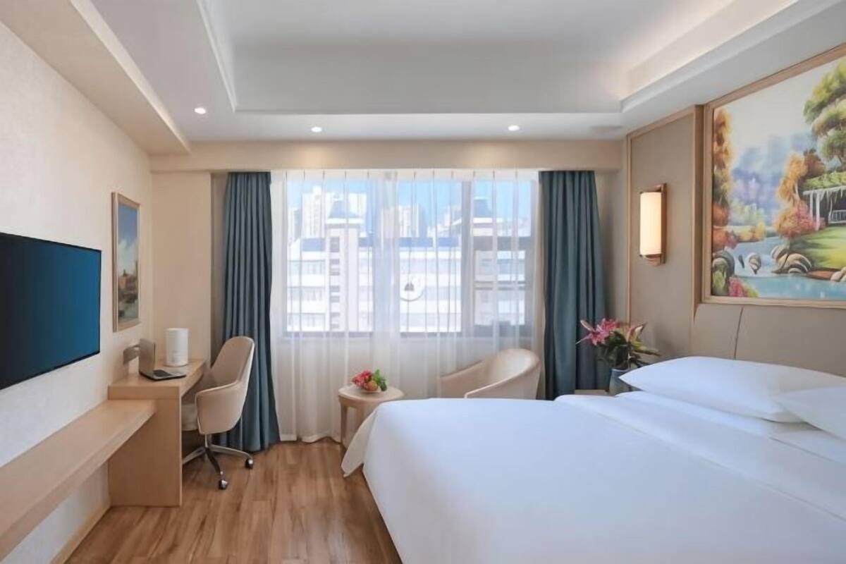 A hotel room with a bed, desk, chair, and a painting on the wall.