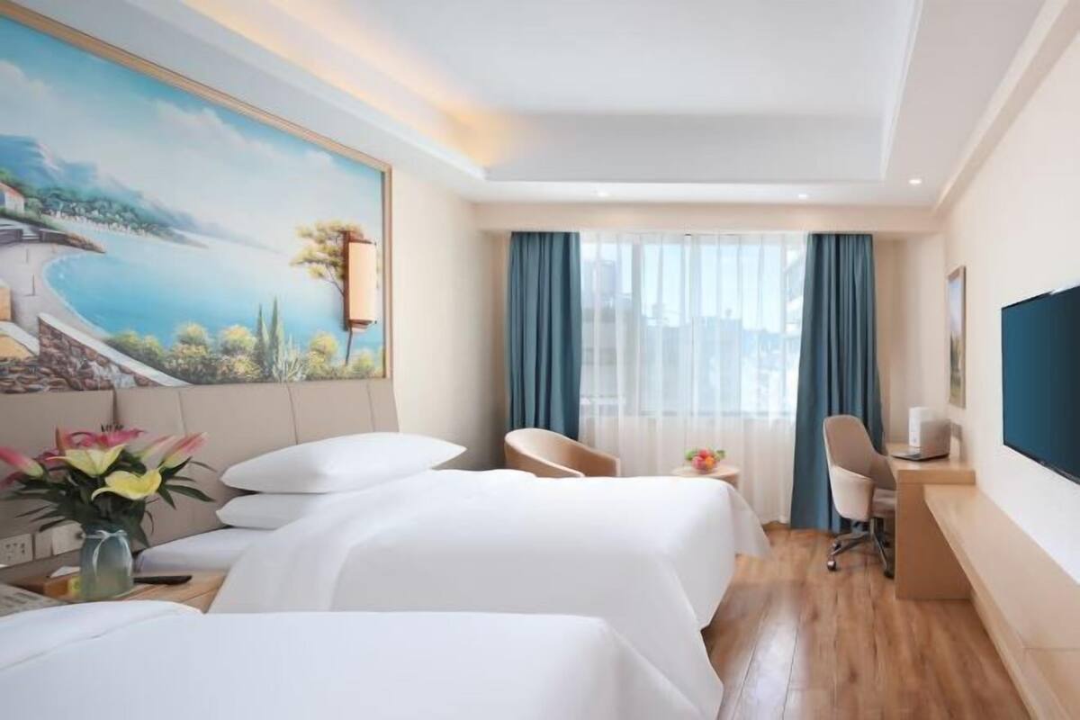 A hotel room with two beds, a desk, a chair, a TV, and a painting of a landscape.