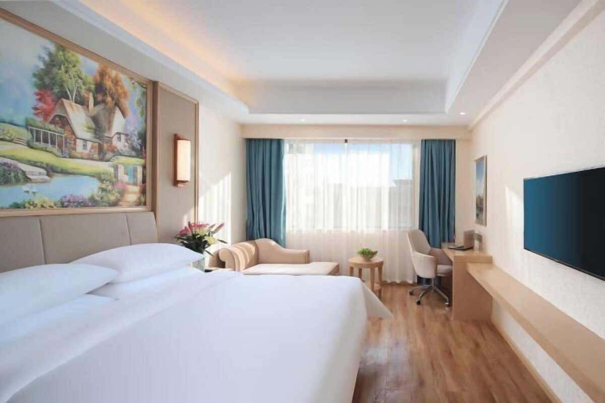 A hotel room with a large bed, a desk, a chair, a television, and a painting on the wall.