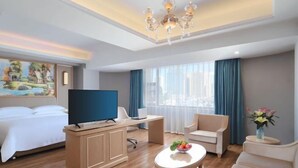 A hotel room with a bed, a desk with a TV, a chair, a round table with flowers, and a large window with curtains. - Vienna Hotel (Kunming Baiyun Road Metro Station Jiang'an) (Kunming)