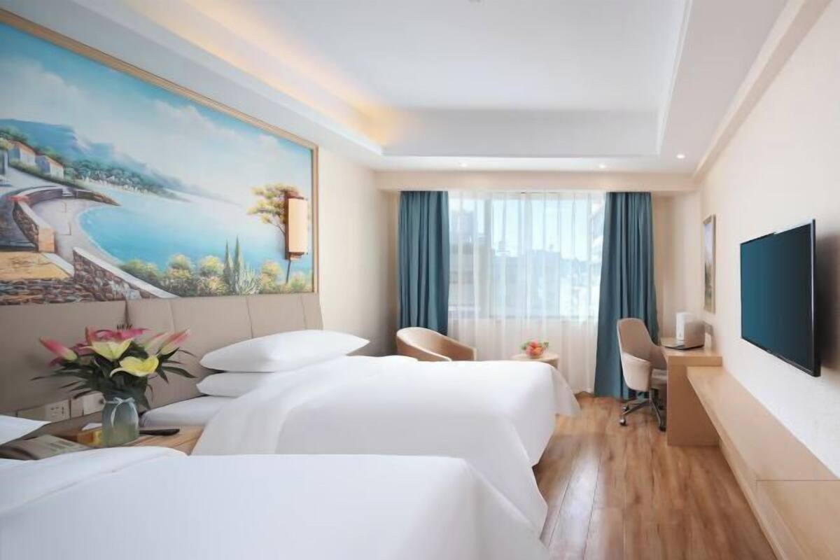 A hotel room with two beds, a desk, a chair, a TV, and a painting of a coastal scene.