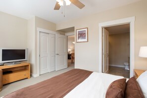 A bedroom with a bed, a TV on a stand, a ceiling fan, and a door leading to a bathroom.