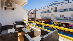 A balcony with wicker furniture, a pool, and buildings with balconies and shops. - 2BD Apartment in Alagoa Beach by LovelyStay (Altura)