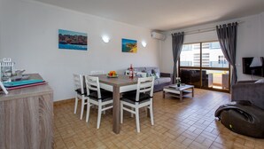A modern living room with a dining area, a sofa, a TV, and a balcony with a view. - 2BD Apartment in Alagoa Beach by LovelyStay (Altura)