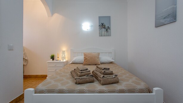 A neatly made bed with folded towels, a bedside table with a lamp, and a wall-mounted picture. - 2BD Apartment in Alagoa Beach by LovelyStay (Altura)