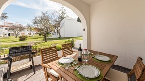 Un espace extérieur aménagé pour les repas, avec une table en bois pour deux personnes, un barbecue et une vue sur une cour intérieure bordée de bâtiments et d’arbres.