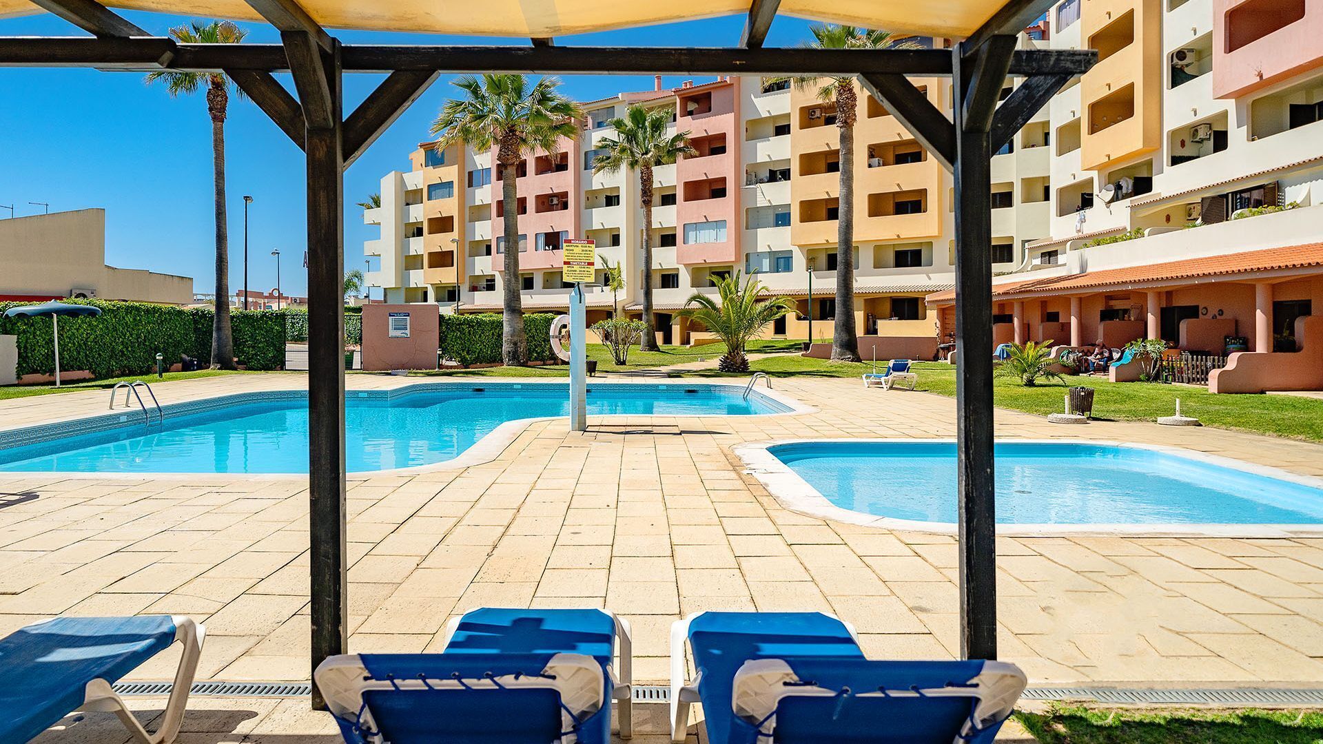 Ett poolområde med två pooler, solstolar och en möblerad sittplats under tak.