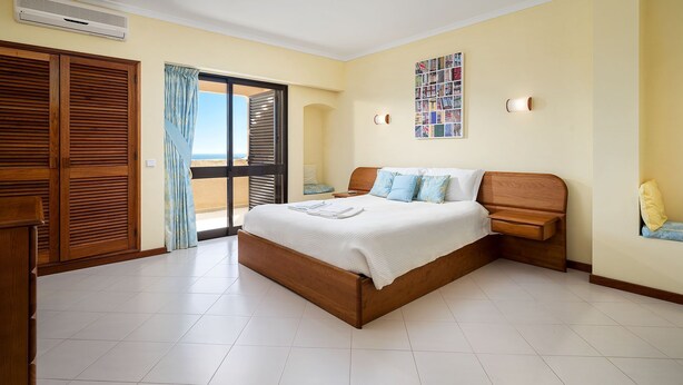 A bedroom with a large bed, wooden headboard, a nightstand, a wall-mounted lamp, and a view of the beach through a sliding door.