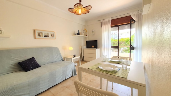 A cozy living room with a sofa, a dining table set for two, a TV, and a window with curtains.