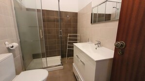 A bathroom with a glass shower enclosure, a toilet, a sink with a mirror, and a wooden door. - Lovely Algarvian Escape by LovelyStay (Manta Rota)