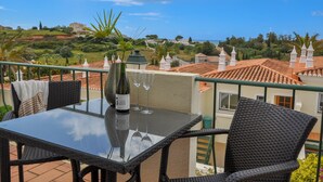 A balcony with a table, chairs, a bottle of wine, and glasses. The balcony overlooks a town with red-roofed buildings and greenery. - 2BR  Duplex Townhouse w/Pool by LovelyStay (Ferragudo)