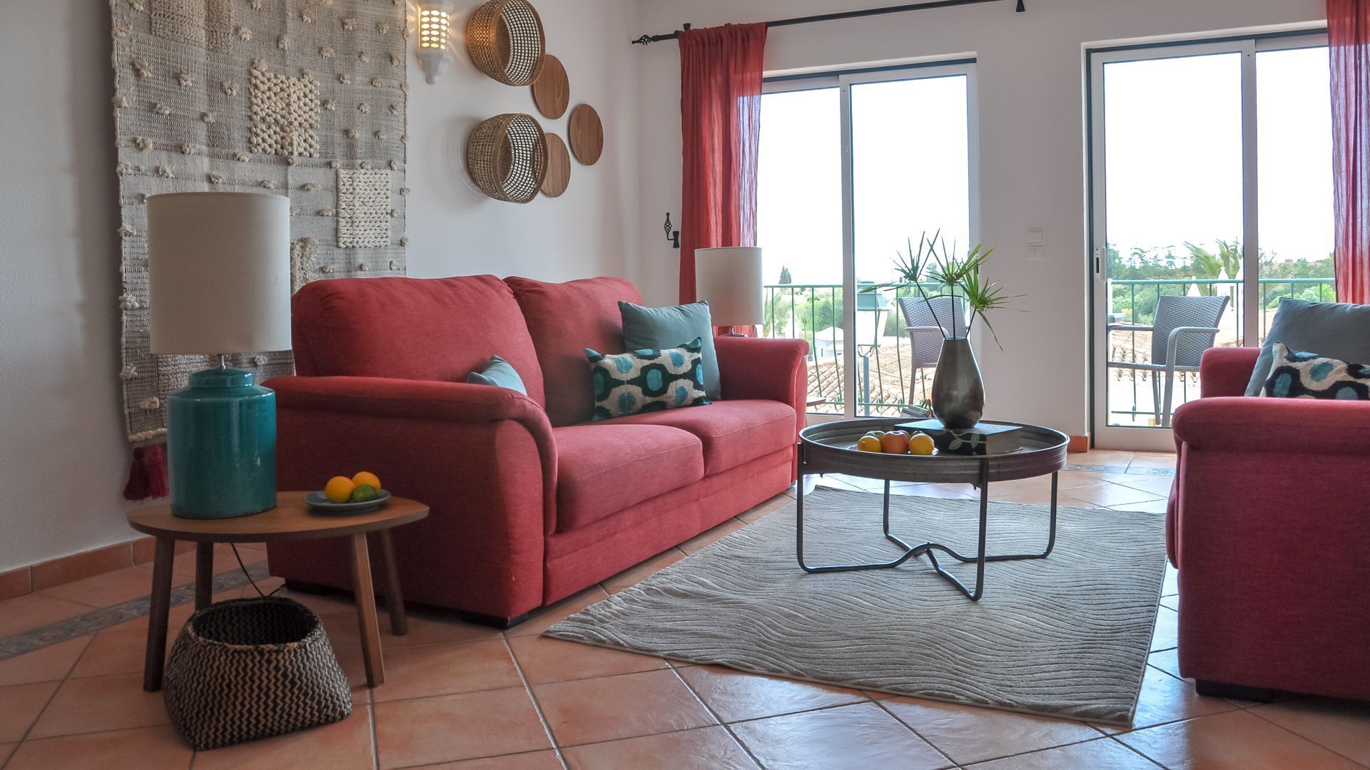 A living room with a red sofa, a coffee table with fruit, a lamp, and a view of the outdoors through large windows.