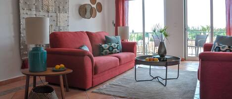 A living room with a red sofa, a coffee table with fruit, a lamp, and a view of the outdoors through large windows.