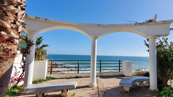 A seaside gazebo with a view of the ocean, a paved walkway, and benches.