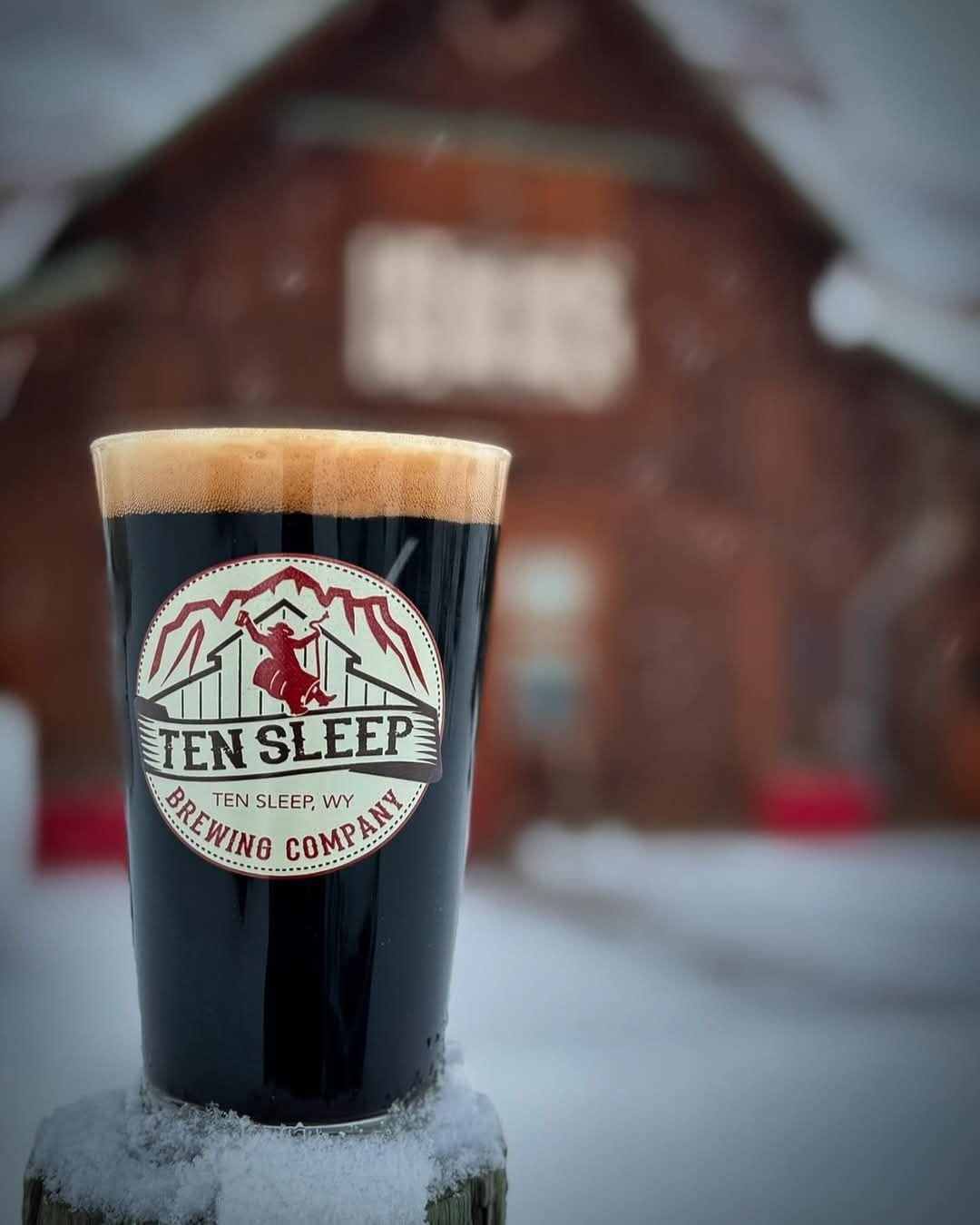 Et glas mørk øl med et logo og teksten "Ten Sleep Brewing Company" foran en træbygning.