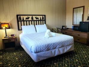 A hotel room with a bed, a nightstand with a lamp, a dresser, a mirror, and a wall with a nature-themed headboard.