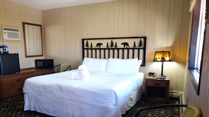 A neatly made bed with white linens, a headboard with a bear and tree silhouette, a bedside lamp, a small table with a radio, and a patterned carpet. - The Carter inn (Ten Sleep)