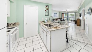 Une cuisine avec des armoires blanches, un comptoir en granit et un évier. On y trouve une porte menant à une autre pièce, un ventilateur de plafond et une vue sur un balcon aménagé avec du mobilier d’extérieur.