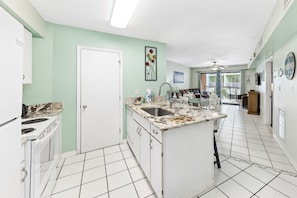 A kitchen with white cabinets, a granite countertop, and a sink. There is a door leading to another room, a ceiling fan, and a view of a balcony with outdoor furniture.