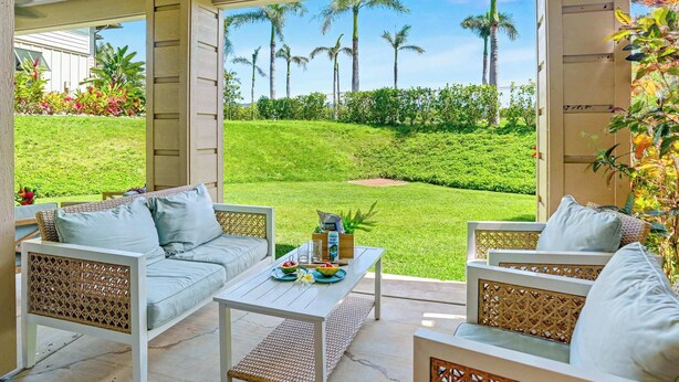 Un área de estar al aire libre con mobiliario de mimbre, una mesa con un bol de fruta y vistas a un césped cuidado y palmeras.