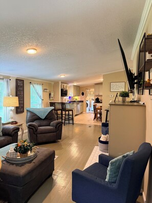 A living room with a sofa, armchair, and ottoman, a kitchen in the background, and a dining area with chairs.