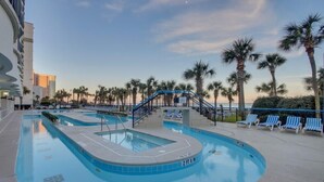 A pool area with a two-tiered pool, lounge chairs, palm trees, and a staircase leading to a deck. - Boardwalk Resort Condo Ocean-front Balcony 🏊 Pool 🏊 (Myrtle Beach)